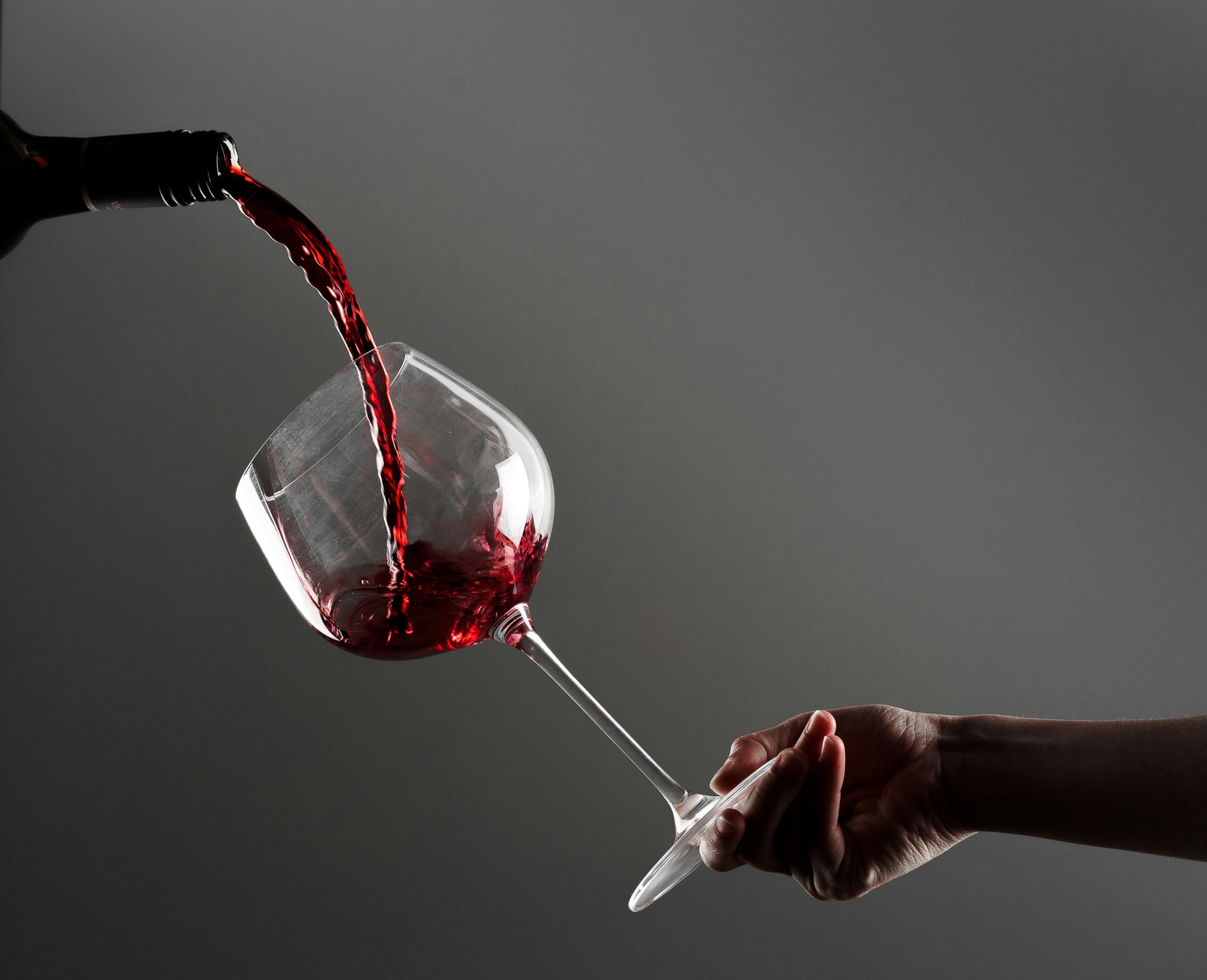 A dramatic, low-key shot of a person's hand holding a large, tilted wine glass as a stream of deep red wine pours from a dark bottle against a grey background.