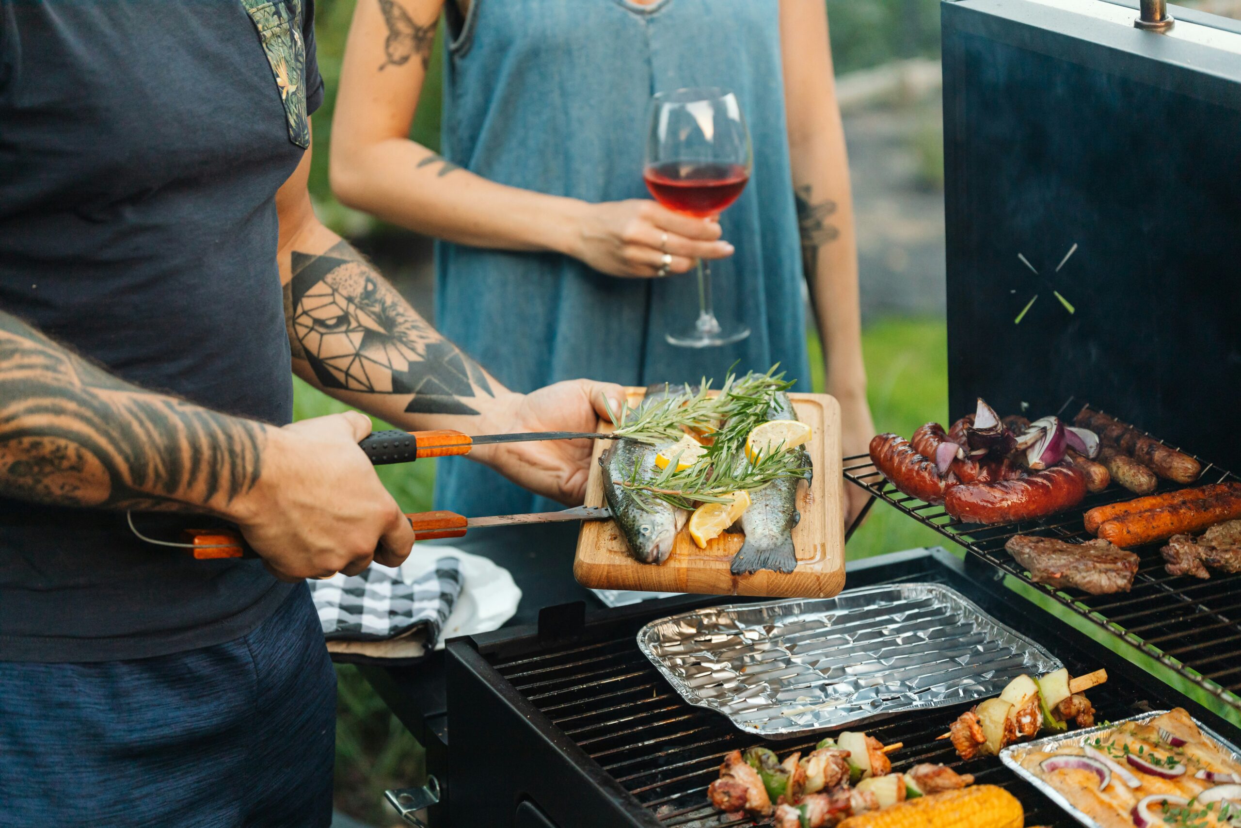 Man with tattooed arms placing rosemary and lemon-stuffed sea bass onto a charcoal barbecue grill alongside sausages, kebabs, and corn, while a woman in a denim dress holds a glass of rosé wine.