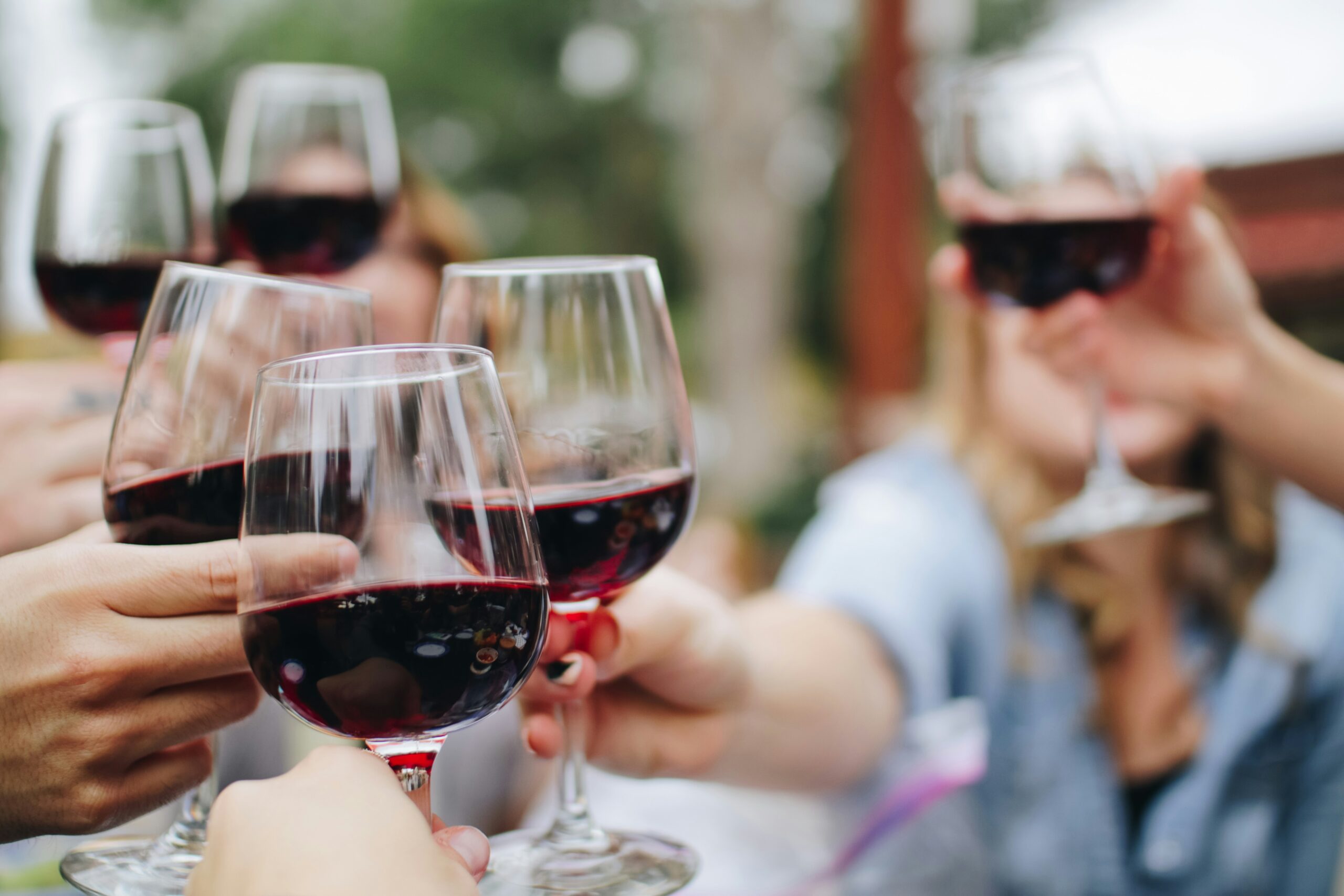 A close-up shot of four people toasting with large, stemmed glasses of red wine at an outdoor gathering, with the background blurred.