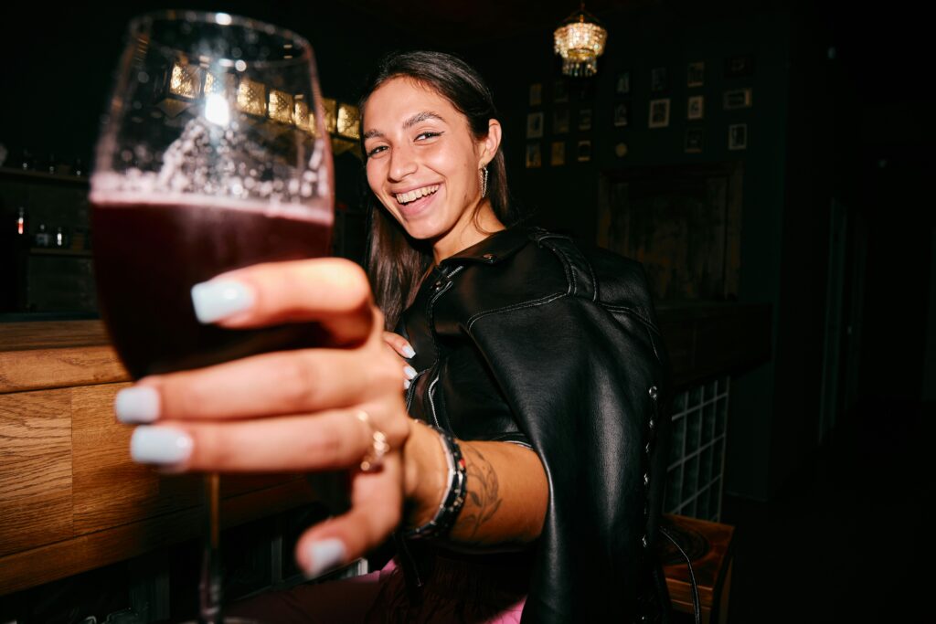 A happy woman in a leather jacket holding up a glass of red liquid in a UK bar, illustrating the choice between traditional vintages and alcohol-free wine for British drinkers.