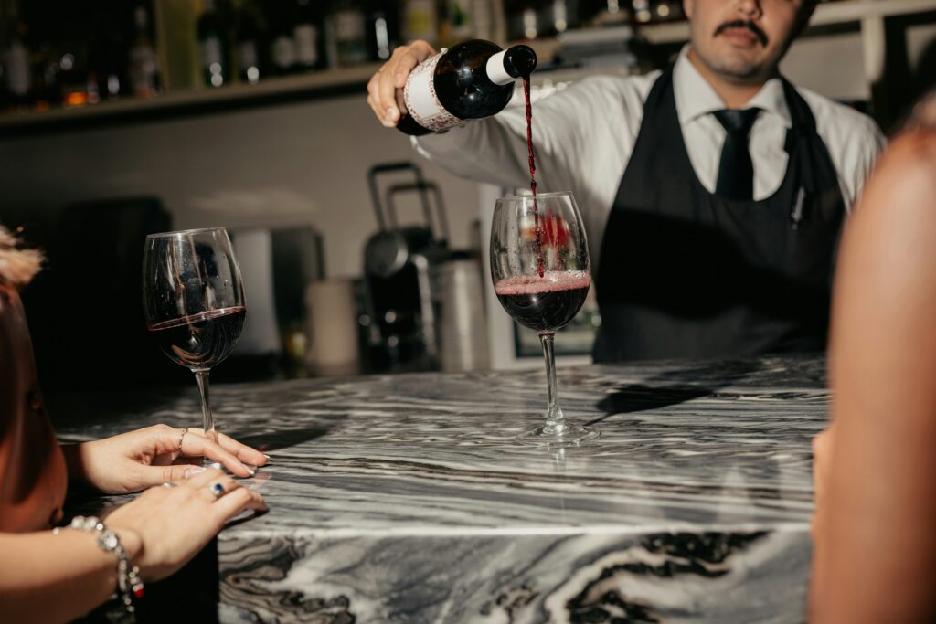 A close-up of red wine being poured from a dark bottle into a standard 750ml glass, visually representing the low liquid value found in a £10 bottle of wine within the 2026 UK market. The scene highlights the minimal amount of actual wine remaining after UK alcohol duty and other taxes are deducted.