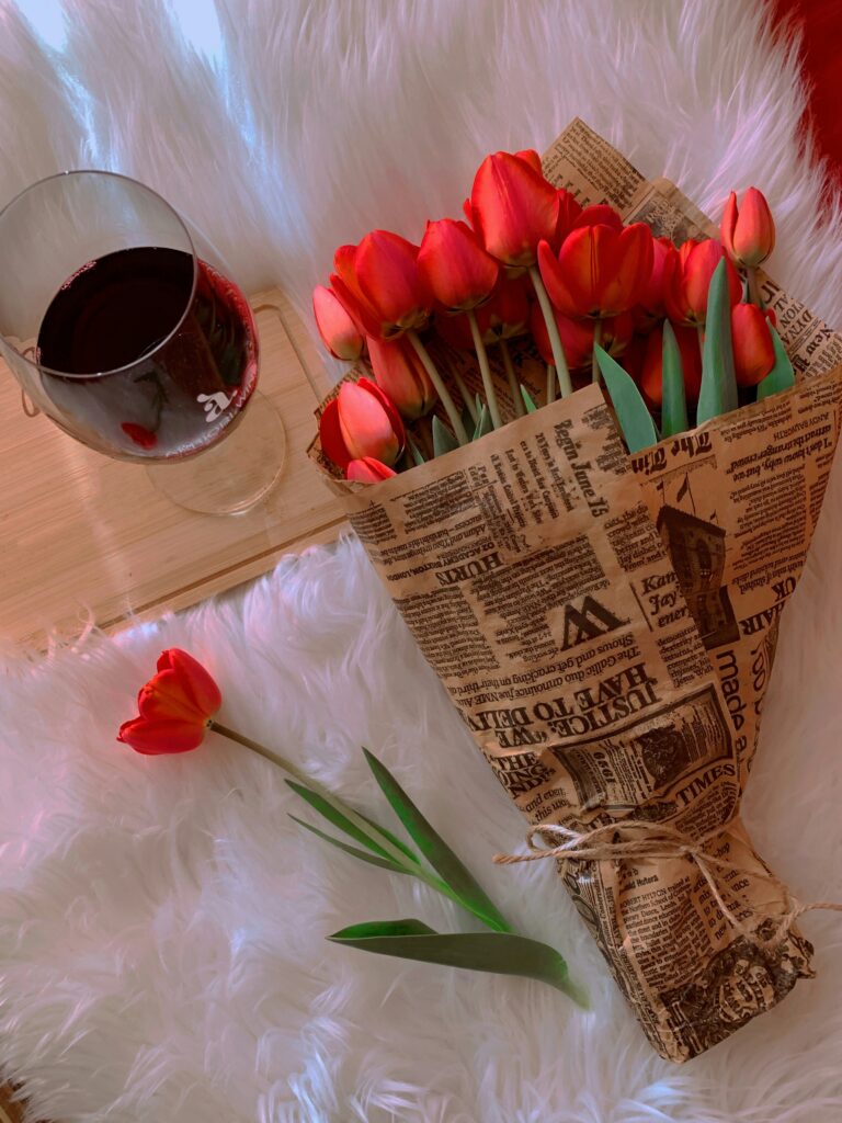 A glass of rich red wine resting on a wooden coaster next to a bouquet of red tulips wrapped in newspaper, creating a romantic setting for Valentine’s Day that avoids expensive Valentine’s Day wine myths.