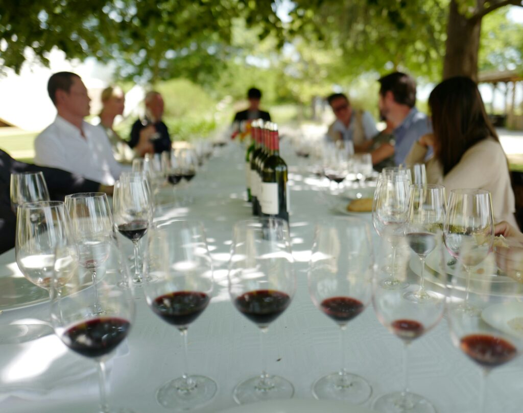 An outdoor wine tasting event in the UK wine market featuring guests seated at a long table, showcasing various wine subscription models and a uk wine subscription experience with multiple glasses of red wine.