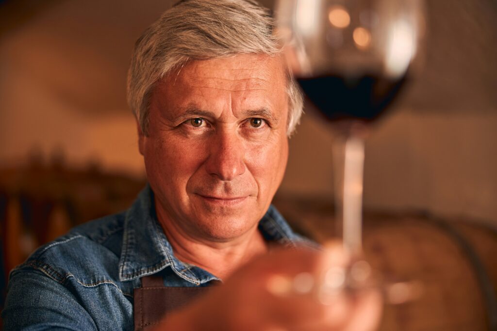 An expert winemaker in a rustic cellar holds up a glass of deep red old vine wines to inspect its clarity and colour as part of his professional wine tasting tips.