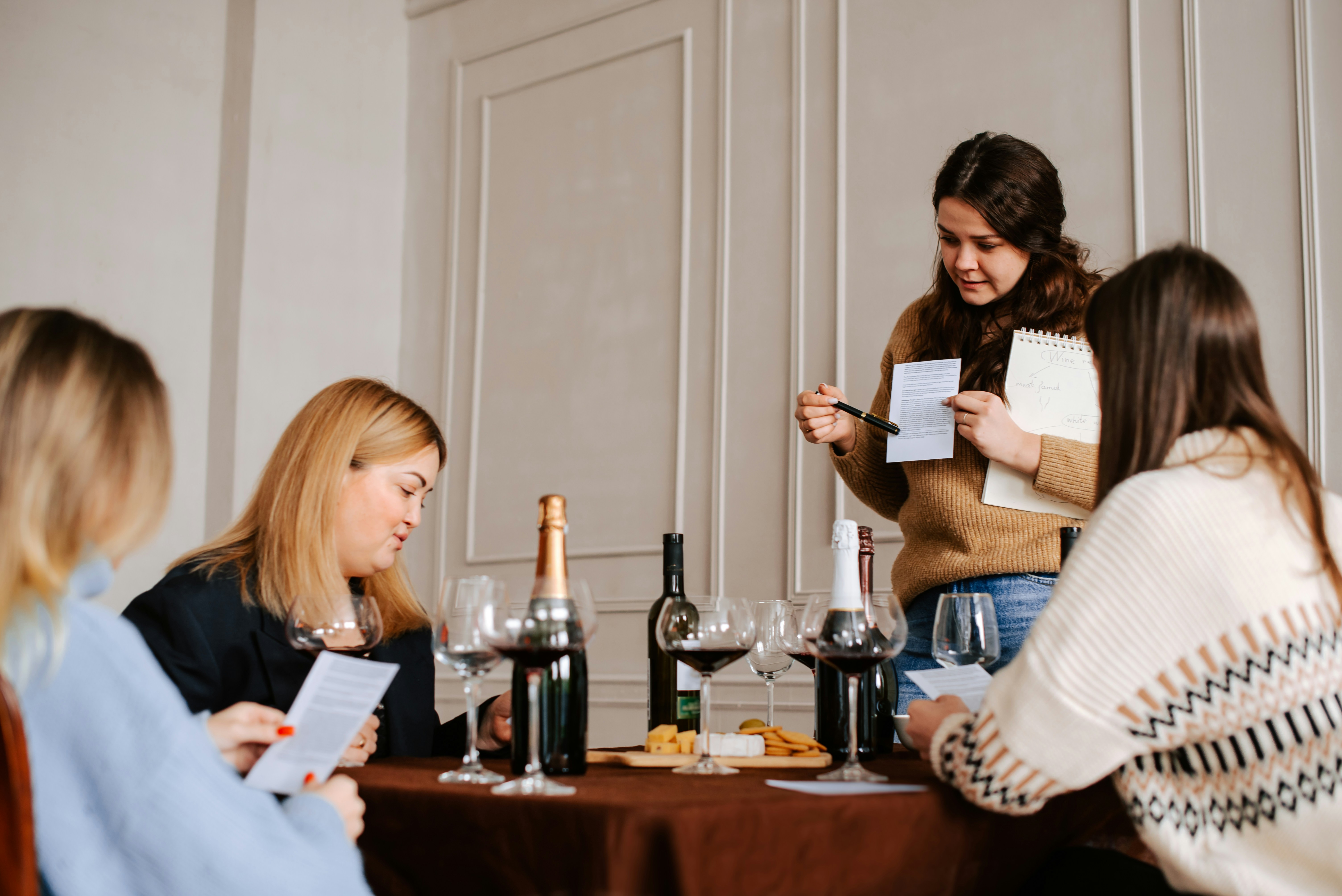 A wine educator standing at a table and showing a group of students how to read wine scores and technical notes on a printed guide.