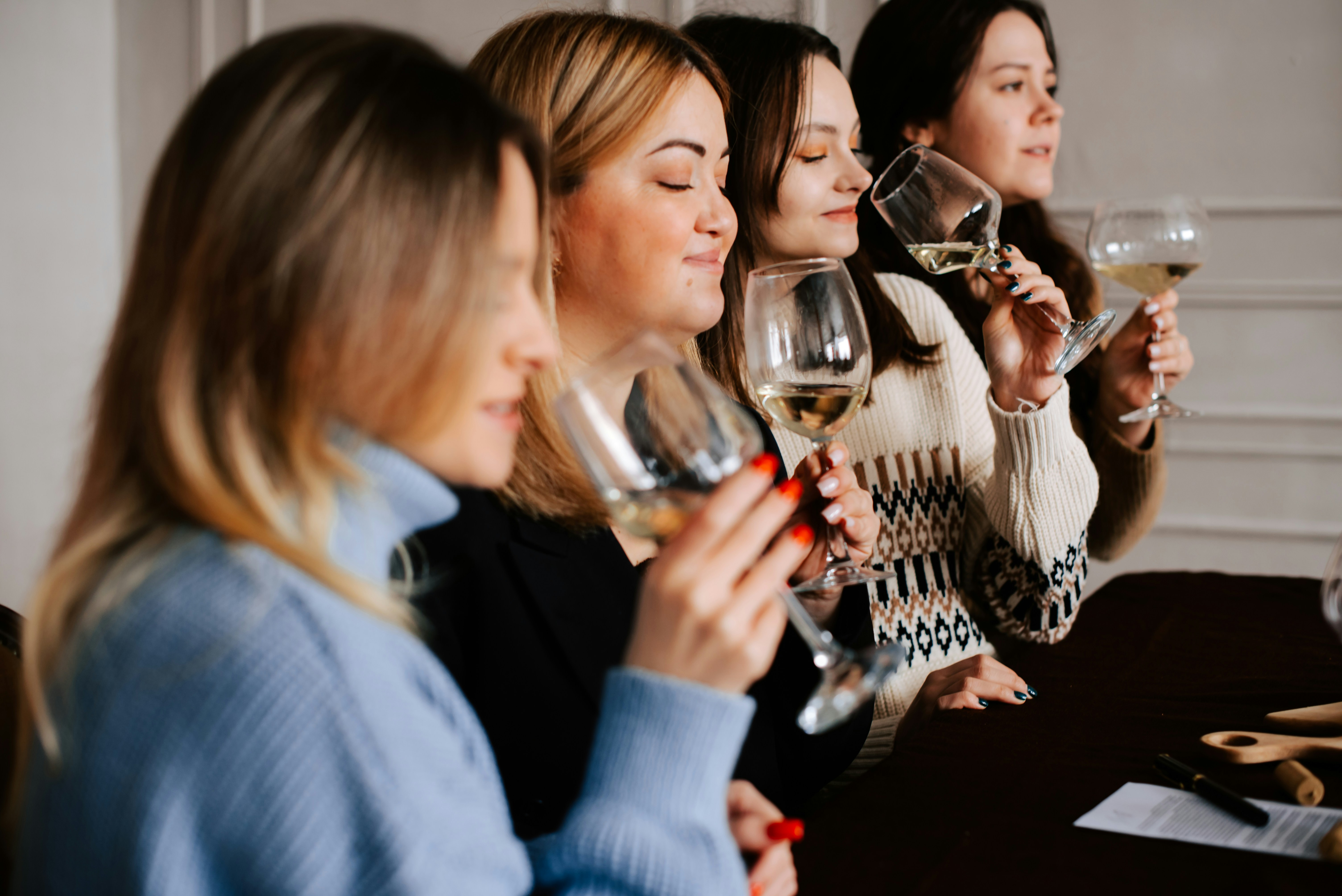 A group of women sitting at a table participating in a professional tasting to have a wine scoring system explained while holding glasses of white wine.